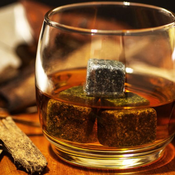 A glass of whiskey on the rocks in simple glass ware with whiskey stones in a carpentry work space with a hammer and saw with wood chips on top a wood desk.